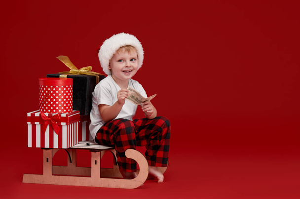 A little boy in a Santa hat sits on a wooden sled with gift boxes and holds a banknote. The concept of Christmas and New Year Cashback. Discounts and sales - Foto, Imagem
