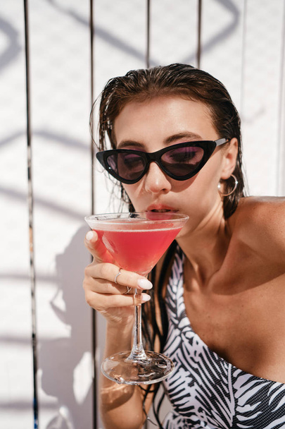 Portrait of attractive brunette girl in one-piece swimsuit and sunglasses thoughtfully drinking cocktail over wood background on beach - Foto, afbeelding