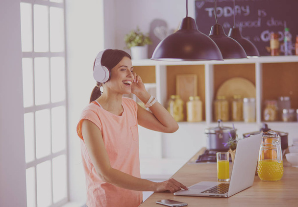 Portrait of a cheerful young woman listening to music with headphones and using laptop computer while standing at the kitchen - Foto, afbeelding