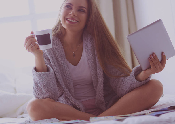 Relaxed young woman sitting on bed with a cup of coffee and digital tablet - Foto, Imagem