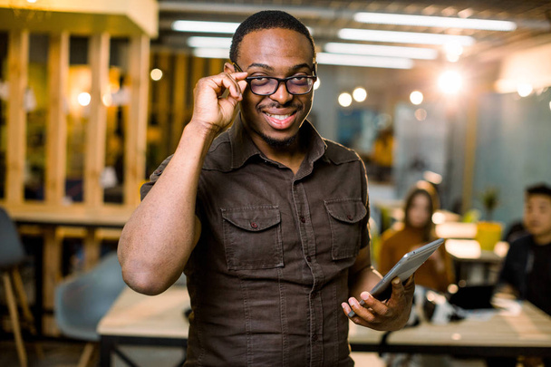 Happy young African man holding digital tablet while standing in creative office. Concept of coworking people. Blurred background with office colleagues on the meeting - 写真・画像