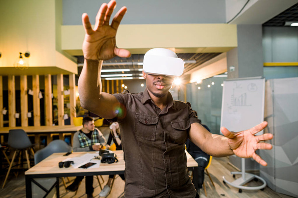 African-American man in casual wear using 3d virtual reality headset. Black gamer wearing goggles, gesturing and having fun in modern coworking place. Colleagues on the background - Photo, image