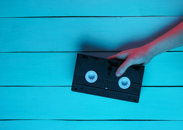 Popcultuur, retro Wave, nachtleven. Vrije tijd. Hand houdt video cassette op blauwe houten achtergrond. Neon rood blauw licht. Retro stijl. Top View - Foto, afbeelding