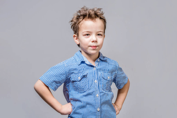 Cute boy holds hands on waist with shy look on gray background.  - Foto, afbeelding