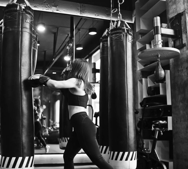 Fittes Mädchen in Sportbekleidung und Boxhandschuhen schlägt in einer dunklen Turnhalle gegen einen Boxsack. Selbstverteidigungstraining für Frauen. die Fähigkeit, sich selbst zu kneifen. schwarz-weiß - Foto, Bild