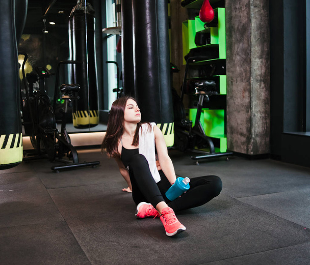 Time for break. Tired fit girl in sportswear sits on the floor and holds bottle of water with towel on her shoulders in the gym. Sports concept - Photo, image