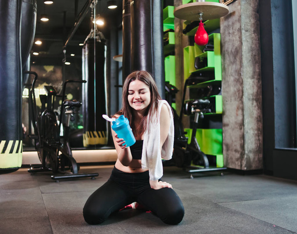 Time for break. Tired fit girl in sportswear sits on the floor and holds bottle of water with towel on her shoulders in the gym. Sports concept - Fotoğraf, Görsel