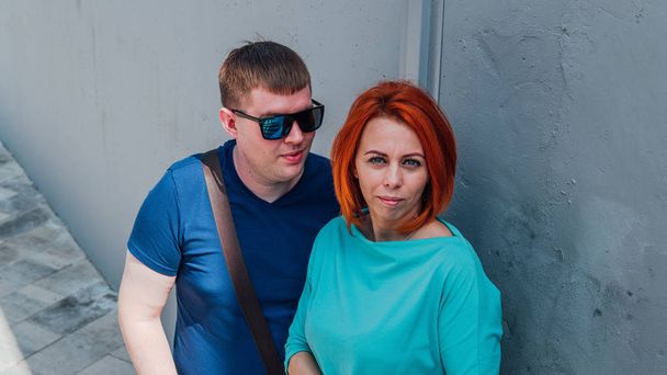 Happy couple in casual clothes are on a walk. Happy man and woman. A man in sunglasses with red-haired woman against a stone wall. - Fotografie, Obrázek