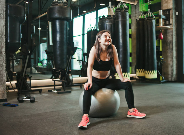 Sportlächelnde Frau in Sportkleidung sitzt auf einem Fitball in einem dunklen Fitnessstudio. Pausenzeit - Foto, Bild