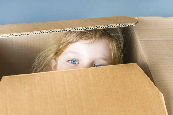 Little blue-eyed  girl looking out of a cardboard box, close-up. - Φωτογραφία, εικόνα