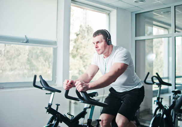 Healthy lifestyle concept. Young sporty man in white t-shirt and shorts is exercising bike at spinning class . Cardio training - Photo, Image