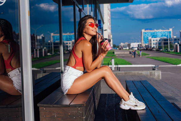 Stylish happy young brunette woman wearing white shorts and sneakers holding pink cup of coffee to go sitting next to coffee shop. - Photo, Image
