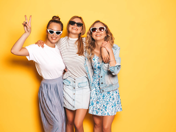 Three young beautiful smiling hipster girls in trendy summer clothes. Sexy carefree women posing near yellow wall in studio. Positive models going crazy and having fun.They show peace sign - 写真・画像