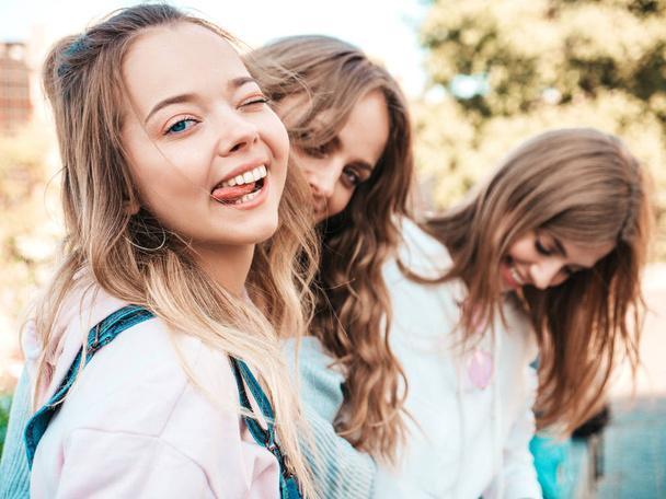 Porträt von drei schönen jungen lächelnden Hipstermädchen in trendiger Sommerkleidung. sexy unbeschwerte Frauen posieren auf der Straße Hintergrund. Positive Modelle mit Spaß. Augenzwinkern und zeigt Zunge - Foto, Bild