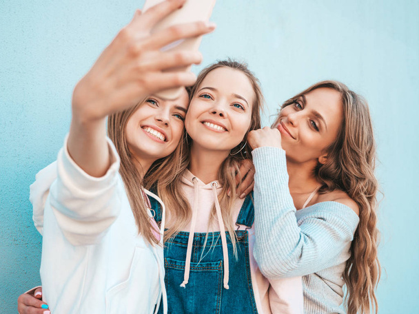 Три улыбающиеся хипстерки в летних одеждах. Девушки, делающие селфи автопортретные фотографии на смартфонах.
 - Фото, изображение