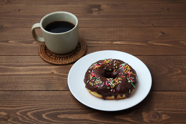 木製のテーブルの上にホットコーヒーとプレート上のチョコレート焼きドーナツ - 写真・画像
