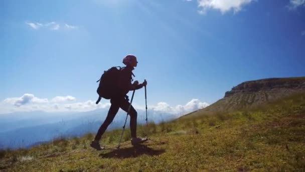 Young woman with backpack and trekking poles walks uphill towards the summit against blue sky. Slow motion. Lady is hiking in beautiful Caucasian mountains. - Footage, Video