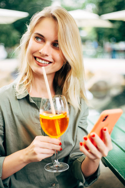 Photo of cheerful blonde with cocktail and phone in her hands on street - Foto, afbeelding