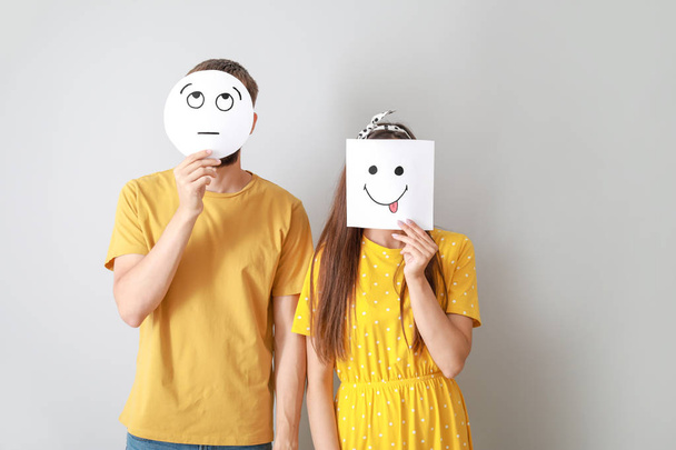 Couple hiding faces behind sheets of paper with drawn emoticons on grey background - Foto, Imagen