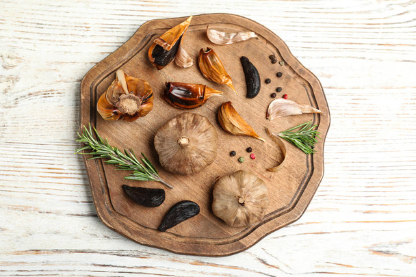 Board with black garlic and rosemary on white wooden table, top view - Foto, afbeelding