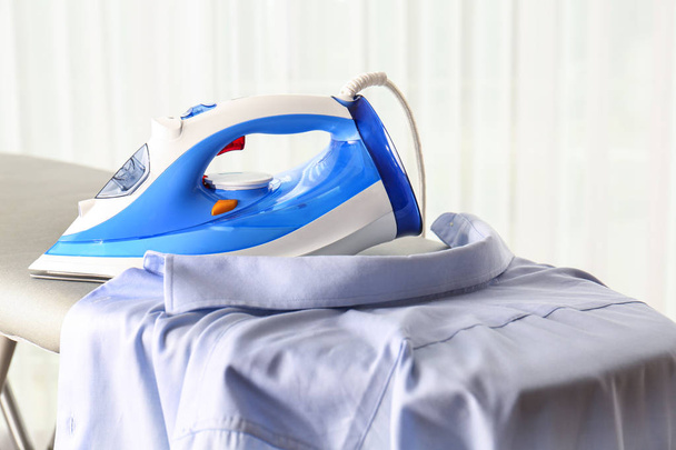 Modern electric iron and clean shirt on board against light background - Foto, Bild