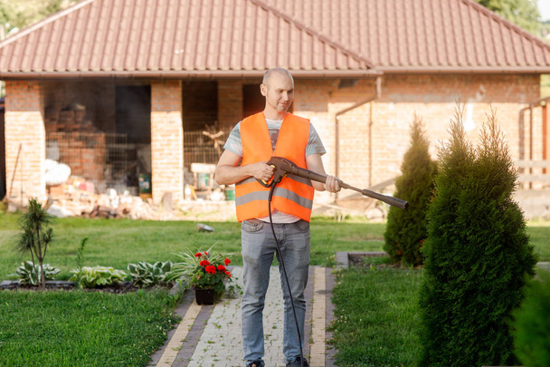 Un uomo con un giubbotto arancione pulisce una tegola d'erba nel suo cortile vicino alla casa. Pulizia ad alta pressione
 - Foto, immagini