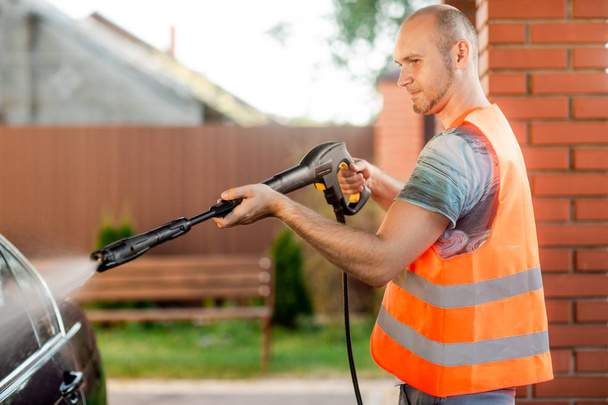 A man in orange vest washes his car with a large head of water from a karcher on open air. High pressure cleaning - Photo, Image