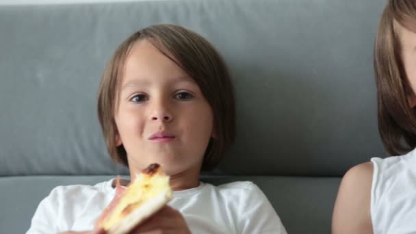 Cute children, sitting on couch, eating pizza and watching TV. Hungry child taking a bite from pizza on a pizza party day at home - Materiał filmowy, wideo