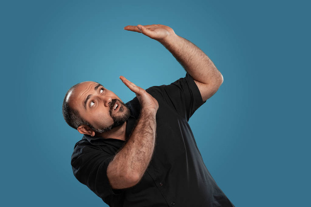 Close-up portrait of a brunet good-looking mature male with beard, dressed in a black t-shirt, looking afraid that something will fall on him while posing against a blue studio background. Sincere emotions concept. Copy space. - Foto, Imagen