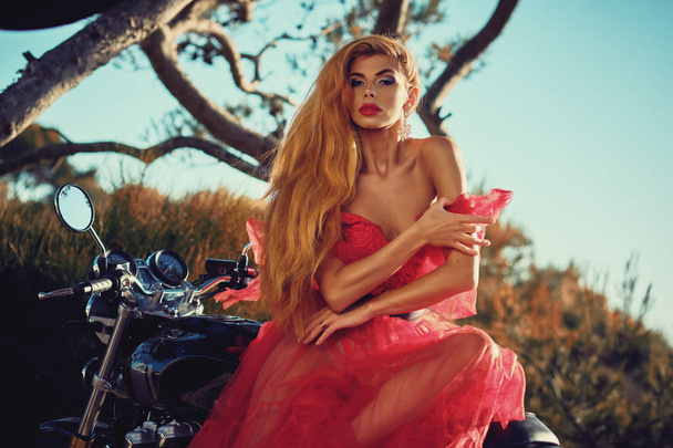 Beautiful woman wearing dress posing seated on motorcycle outdoors - Zdjęcie, obraz