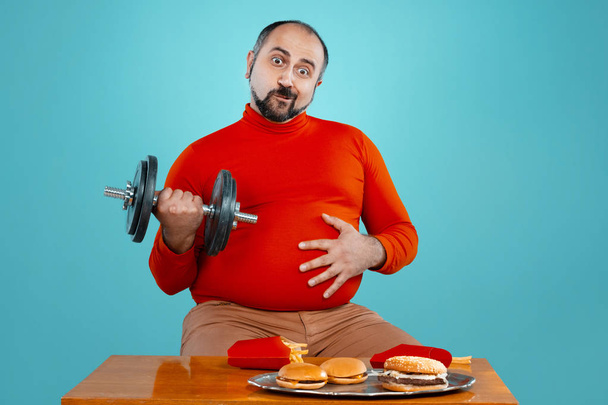 Close-up portrait of a brunet middle-aged guy with beard, dressed in a red turtleneck, looking at the camera while posing with dumbbell at the table with burgers and french fries against a blue studio background. Sincere emotions concept. Copy space. - Photo, Image