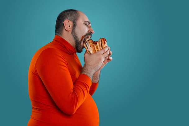 Close-up portrait of a brunet stately mature man with beard, dressed in a red turtleneck, eating burger while posing sideways with against a blue studio background. Sincere emotions concept. Copy space. Fast food. - Foto, imagen