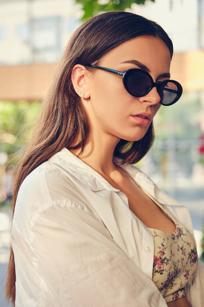 Porträt eines Mädchens mit dunkler Sonnenbrille, das in der Stadt nahe der Sommerterrasse posiert. Bekleidet mit Oberteil mit Blumenmuster, weißem Hemd, schwarzer Hose, Taille. - Foto, Bild