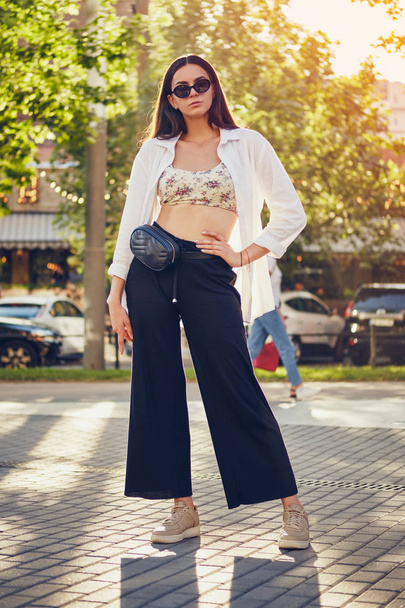 Portrait of a girl in dark sunglasses posing in city. Dressed in top with floral print, white shirt, black trousers, waist bag and sneakers. - Fotoğraf, Görsel