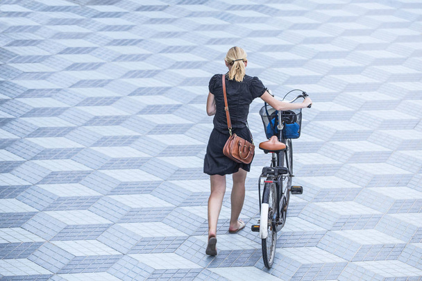 Jovem caucasiana com aspecto urbano caminha com sua bicicleta através de uma área solitária
 - Foto, Imagem