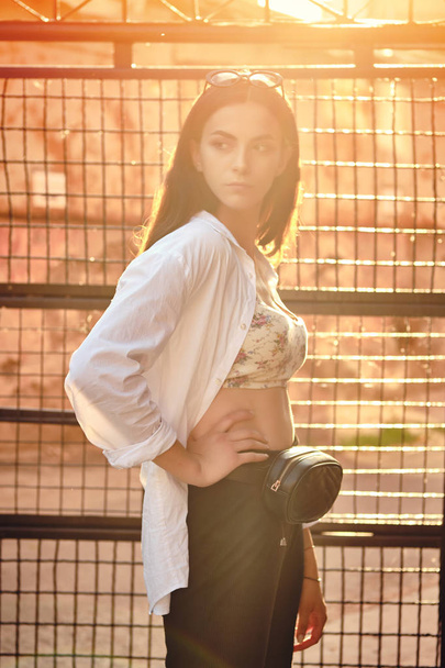 Portrait d'une fille aux lunettes de soleil sombres posant en ville contre une clôture à treillis. Habillé en haut avec imprimé floral, chemise blanche, pantalon noir, sac de taille
. - Photo, image
