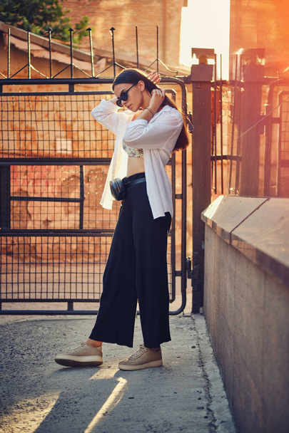 Portrait of a girl in dark sunglasses posing in city. Dressed in top with floral print, white shirt, black trousers, waist bag and sneakers. - Foto, imagen