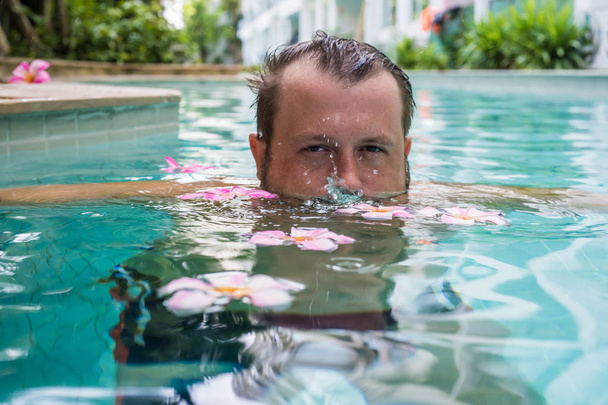 задумчивый бородатый мужчина держит цветы в бассейне. Тропические цветы Frangipani Plumeria, Leelawadee плавающие в воде. SPA бассейн. Мир и спокойствие. Концепция
. - Фото, изображение