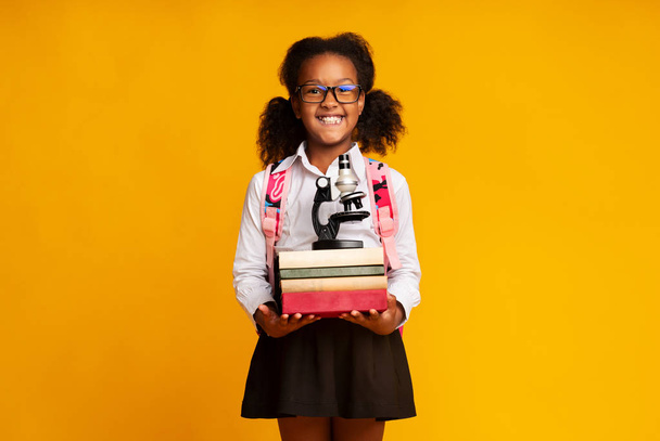 Afro Schoolgirl Holding Microscope And Stack Of Books, Yellow Background - Φωτογραφία, εικόνα