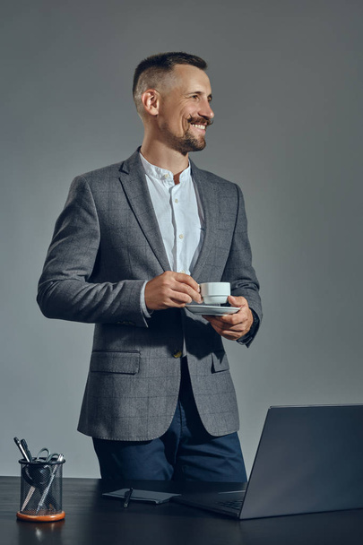 Vousatý podnikatel se stylovým knírem, oblečený v klasickém obleku pracuje na notebooku, zatímco sedí u stolu v kanceláři, šedé pozadí. - Fotografie, Obrázek