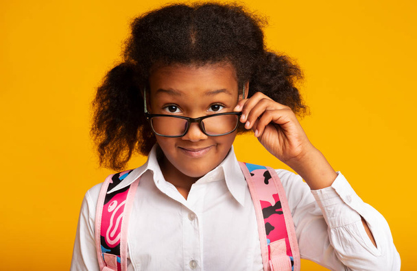 Elemental estudiante chica mirando por encima de gafas en cámara, fondo amarillo
 - Foto, Imagen