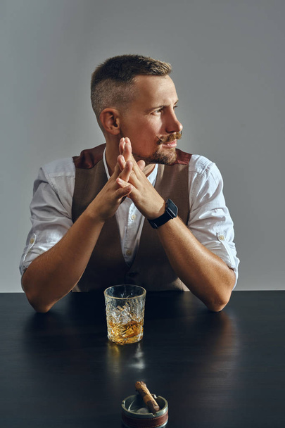 Man met stijlvolle snor, gekleed in klassiek bruin vest, wit shirt zit aan tafel, genieten van whisky, sigaar. Grijze achtergrond, close-up foto. - Foto, afbeelding