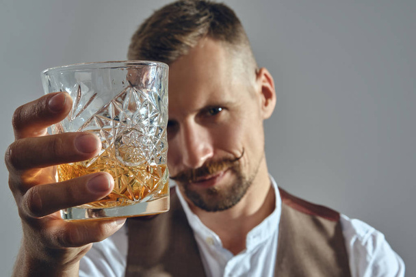Man with stylish mustache, dressed in classic brown vest, white shirt is sitting at the table, enjoying whiskey. Grey background, close-up shot. - Photo, image