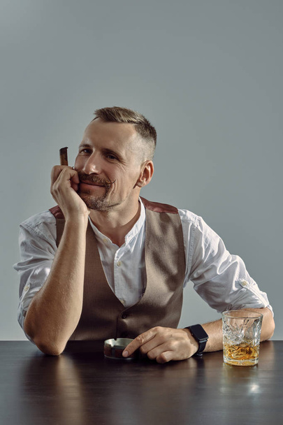 Man with stylish mustache, dressed in classic brown vest, white shirt is sitting at table, enjoying whiskey, cigar. Grey background, close-up shot. - Fotó, kép
