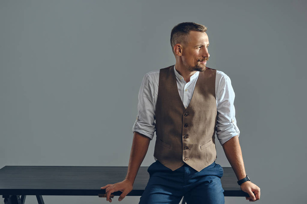 Man with a stylish mustache, dressed in brown vest, white shirt and dark trousers is posing sitting on table. Grey background, close-up. - Photo, Image