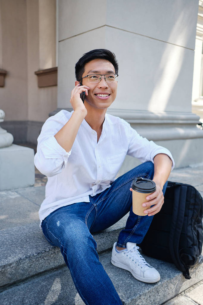 Joven alegre asiático macho estudiante en gafas con mochila felizmente hablando en celular cerca de la universidad
 - Foto, imagen