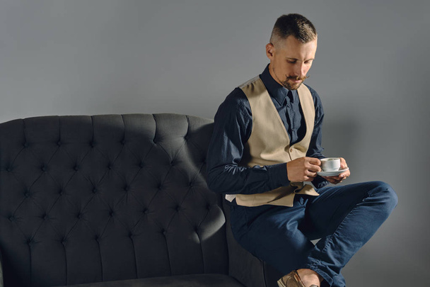 Man with stylish mustache, dressed in black shirt and trousers, beige vest is sitting on dark sofa, holding cup of coffee. Grey background, close-up. - Photo, Image