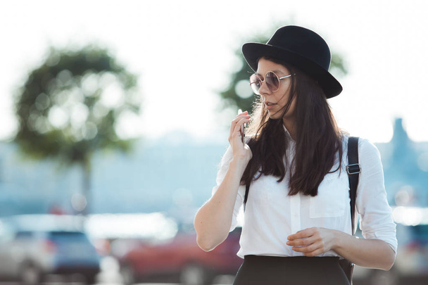 young woman in a hat and with a backpack walks in the city and uses a smartphone. - 写真・画像