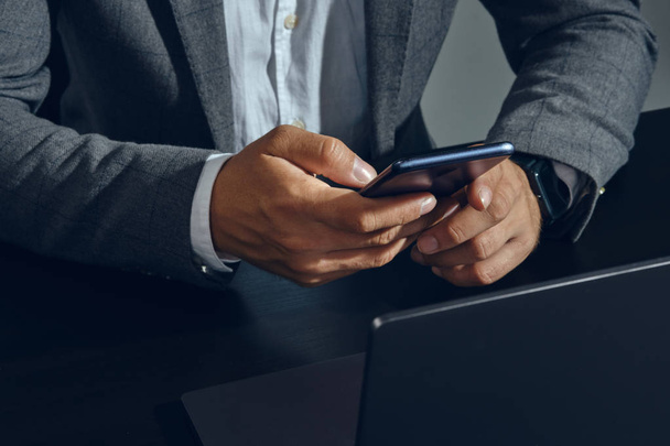 Hands of businessman, dressed in classic suit, white shirt, posing sitting at the table in office, using his smartphone, gray background. Close-up. - Photo, image