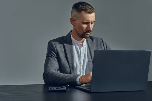 Baard zakenman met stijlvolle snor, gekleed in een klassiek pak werkt op laptop terwijl zittend aan tafel in kantoor, grijze achtergrond. - Foto, afbeelding